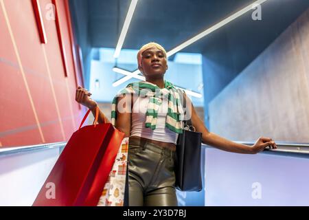 Low angle view portrait of a stylish african woman with short hair coming down an escalator carrying shopping bags Stock Photo