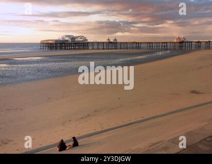 Two women watching the sun setting over the historic north pier in blackpool with glowing light reflected on the beach and colourful twilight sky Stock Photo