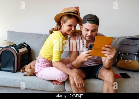 Joyful young couple sitting on their sofa at home, booking their travel online and excitedly preparing for their summer vacation. Stock Photo