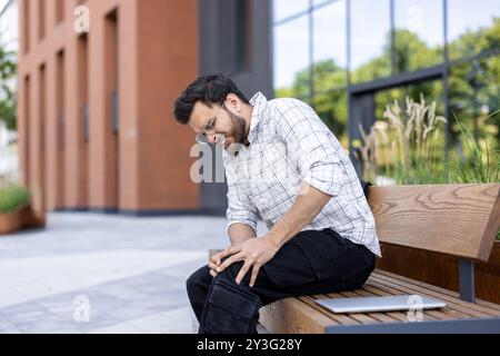 A young Indian man sits outside a building on a bench with his hands on his knees. he feels severe pain in his leg, winced from the injury. Stock Photo