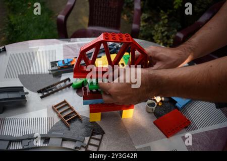 A man assembling toy  bricks on a garden table with colourful tablecloth in a garden. Stock Photo