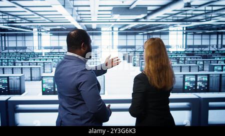 Data center engineer presenting equipment to inspector supervising operations. IT specialist in server farm doing tour for supervisor monitoring equipment performance, camera B Stock Photo