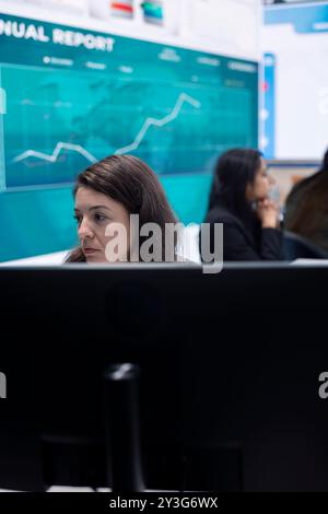 Marketing specialist studying infographics on computer in a monitoring room with big screen, check customer satisfaction from annual data reports. Employee review performance metrics. Stock Photo