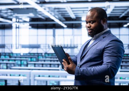 System administrator using tablet in data center with advanced tech, ensuring cybersecurity and efficient network management. Man in server hub optimizing data center equipment with device Stock Photo