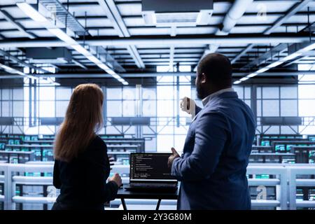 Manager overseeing data center next to employee, doing yearly infrastructure inspection. Coworkers doing brainstorming in server farm, monitoring supercomputers equipment with laptop Stock Photo