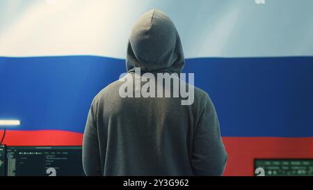 A Russian hacker working in a governmental hacking room with the Russian flag on big screen. Inflicting cyber terror, propaganda, fake news and misinformation in hybrid warfare. Camera A. Stock Photo