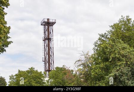 A photo of a tall industrial metal tower with a rusty texture, visible from afar in the middle of trees Stock Photo