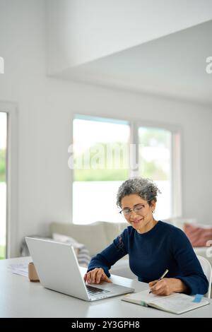 Happy middle aged woman sitting at table and using laptop writing notes. Stock Photo