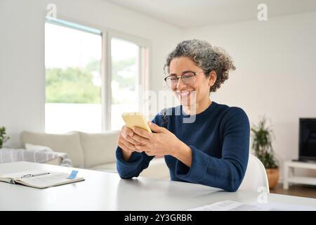 Happy older woman using smartphone sitting at home table in living room. Stock Photo