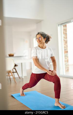 Sporty fit middle aged woman standing on mat at home doing fitness exercises. Stock Photo