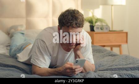 Caucasian man lying on bed using mobile phone smartphone cellphone texting smiling guy male communication online web technology internet social media Stock Photo