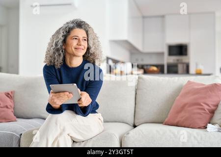 Smiling middle aged woman looking away holding tab relaxing on sofa at home. Stock Photo