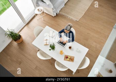 Middle aged woman sitting at table and using laptop at home. Top view. Stock Photo