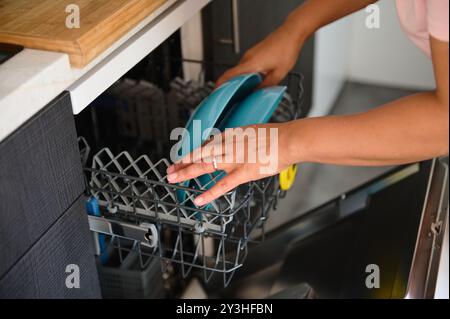 A person places blue plates in an open dishwasher, illustrating daily chores and kitchen organization. Focus on cleanliness and convenience in a moder Stock Photo