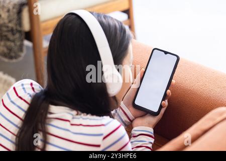 At home, wearing headphones, young asian woman using smartphone while sitting on couch, copy space Stock Photo