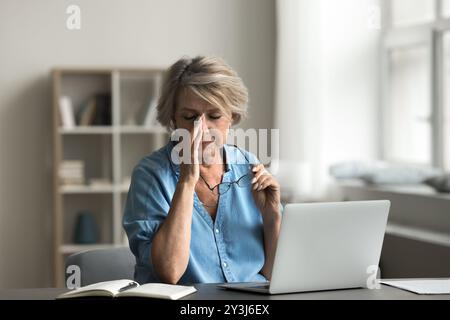 Woman taking off her eyeglasses seated at desk with laptop Stock Photo
