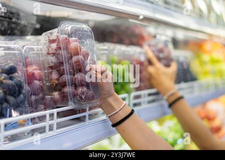 A person selecting a container of grapes from a refrigerated display in a grocery store. Various fruits are visible in the background, showcasing a co Stock Photo