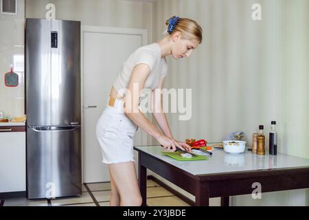 A young woman is chopping onion on a green cutting board in a modern kitchen. The sunlight illuminates the room, highlighting her focused expression a Stock Photo