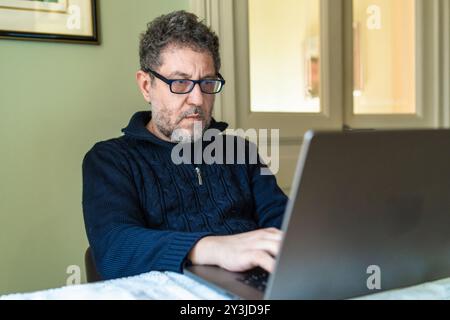 Senior man with glasses working on laptop at home. Mature professional engaged in remote work, showcasing digital literacy and modern work lifestyle f Stock Photo
