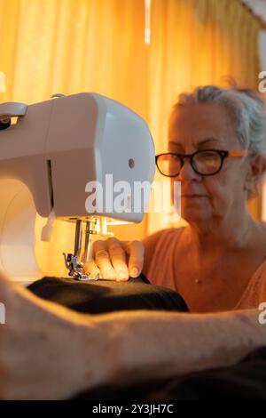 An elderly woman concentrates deeply as she sews a piece of fabric using a sewing machine in a warm-lit room. Stock Photo