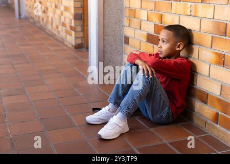 Sitting on floor, boy in school hallway looking away thoughtfully Stock Photo