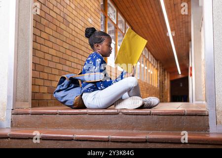 In school, african american girl sitting on steps reading yellow folder with backpack nearby Stock Photo
