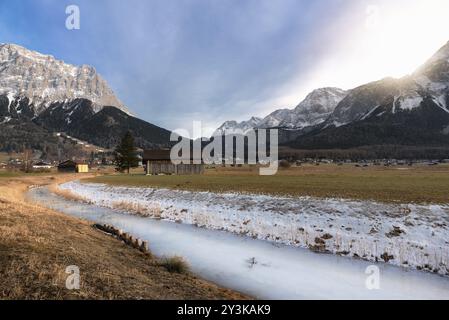 Frozen river on a meadow with dried grass, wooden cabins, a village, and the snow-capped Alps mountains, near Ehrwald, Austria, in December, Europe Stock Photo