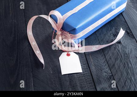 Chic present, wrapped in blue paper and tied with pink ribbon and bow, with an empty label attached to it, on a black wooden table Stock Photo