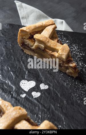 Apple pie slice on a black cutting board decorated with powdered sugar heart shapes, on a gray table. Ready to eat sweets. Traditional dessert Stock Photo