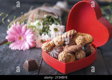 Valentine day theme image with a heart-shaped gift box full of cookies and chocolates, a lovely bouquet of flowers in the background, on a black table Stock Photo