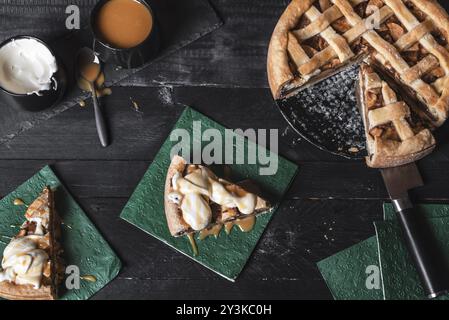 Above view of a rustic table with a sliced apple pie, and two pieces on napkins with cream and caramel sauce. Homemade cake. Traditional classic pie Stock Photo
