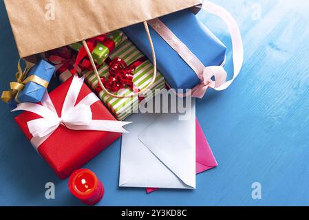 Brown paper bag overturned on a blue wooden table, while multicolored presents and closed envelopes come out of it, near a red, lit candle Stock Photo