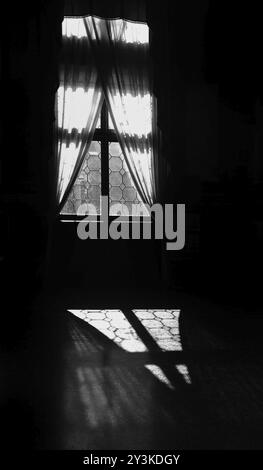 Monochrome atmospheric image of an old window with sunlight shining through closed curtains reflected in shadow on the floor Stock Photo