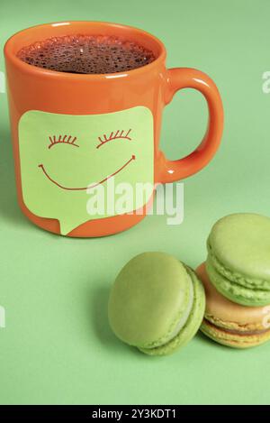 Fresh coffee in an orange mug and a sticky memo with a smiley face beside a stack of colorful macaroons Stock Photo