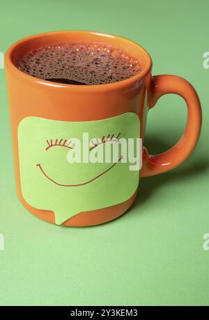 Hot cup of coffee with a sticky paper note on it. Drawn smiley face as a memo on a green sticky note fixed on an orange cup of coffee Stock Photo