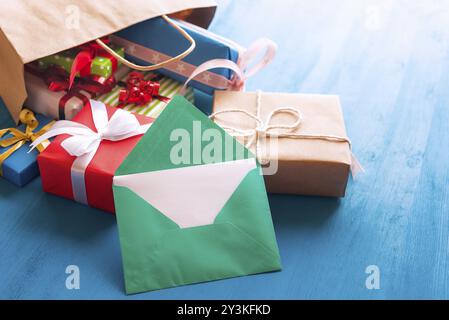 Green envelope with blank white paper leaning against a bunch of colorful presents, that come out of a brown shopping bag, on a blue wooden background Stock Photo