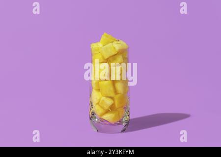 Pineapple pieces in a glass, in bright light, isolated on a purple background. Glass with pineapple pieces minimalist on a purple table Stock Photo