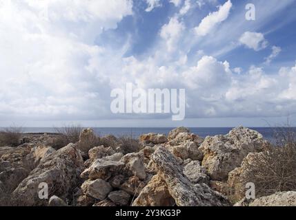 Rough rocks and boulders with dry summer vegetation in front of a calm sea with white sunlit clouds filling the sky Stock Photo