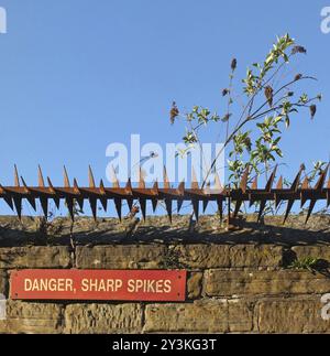 Rusty iron sharp spikes on the top of an old stone wall with a red danger sign against a blue sky Stock Photo