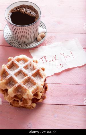 Cute thank you note on a piece of paper with a smiley face on it, on a pink background, near a hot cup of coffee and fresh waffles, with powdered suga Stock Photo