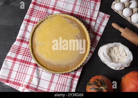 Kitchen table with a pumpkin pie in a tray on a towel surrounded by ingredients, eggs, flour, and pumpkin. Flat lay. Holiday dessert preparation Stock Photo