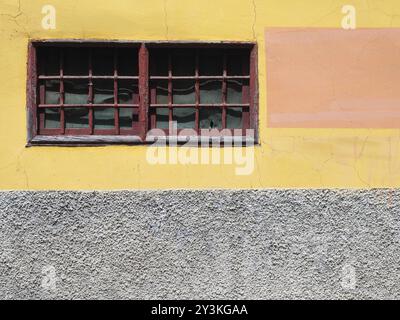 Old painted yellow and grey concrete wall with narrow red framed window with bars and distressed cracked texture Stock Photo