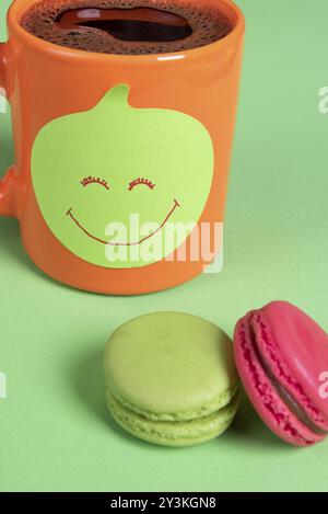 Cup of coffee with a sticky note on it and colorful macaroons on a green table. Smiley face drawn on the paper note Stock Photo