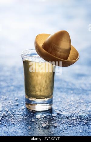 Shot of tequila with salt on a blue table with backlight. Glass of gold tequila with a small sombrero hat. Mexican traditional drink. Strong alcohol Stock Photo
