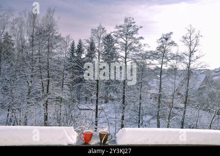 Two cups of hot coffee on a snow-covered railing from a balcony with the snowy trees in the background, on a sunny day of December Stock Photo