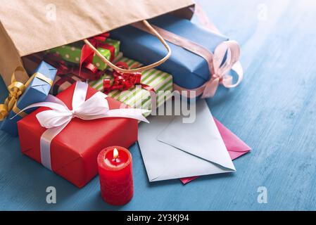 Shopping bag full of colorful presents, paper wrapped and tied with ribbon and bows, overturned on a blue wooden table, over closed envelopes, near a Stock Photo