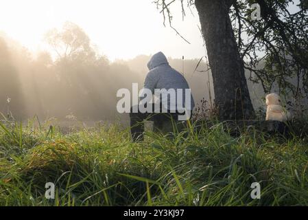 Relationship issues image with a man and a teddy bear sitting on a wooden bench under a tree, while enjoying the morning mist and sun rays coming thro Stock Photo