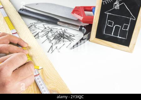 Man's hands measuring a wooden board, having his tools in the background and a funny sketch of a house on a blackboard. Concept about building your dr Stock Photo