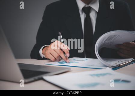 businessman in a suit analyzing documents at a desk. Hand holding pen, pointing at charts. Professional, focused on financial analysis. Stock Photo