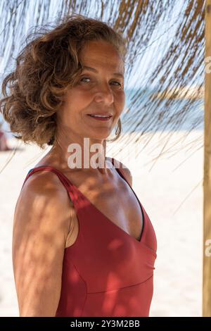 An elderly woman is seen standing on a sandy beach, wearing a red sleeveless top. Stock Photo